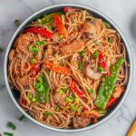 A white bowl of chicken lo mein garnished with green onions and sesame seeds.