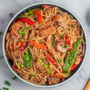 A white bowl of chicken lo mein garnished with green onions and sesame seeds.