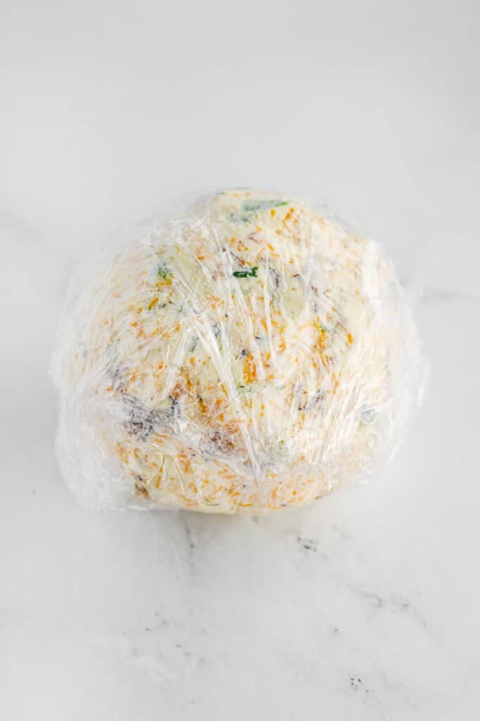 Ball of seasoned cream cheese wrapped in plastic wrap on a marble surface.