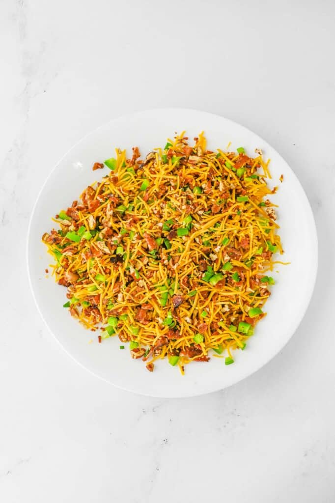 A white plate filled with shredded cheddar cheese, sliced green onions and crumbled bacon on a white background.