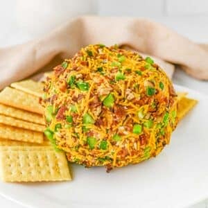 A jalapeno popper cheese ball coated with shredded cheddar cheese, chopped jalapenos and bacon bits, surrounded by rectangle crackers on a white plate.