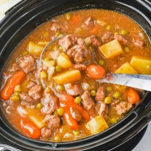 A close-up of a slow cooker filled with beef stew, featuring chunks of beef, potatoes, carrots, peas, and onions in a rich broth.