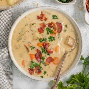 A bowl of creamy New England clam chowder garnished with chopped herbs and crispy bacon bits. A spoon is placed inside the bowl, and a checkered napkin lies next to the bowl on a light-colored surface.