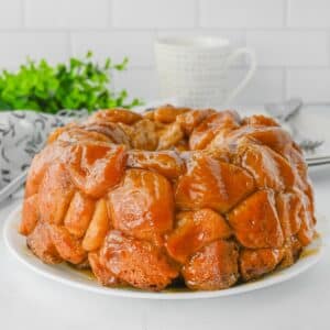 A glazed monkey bread on a white plate. A white mug and green plant are in the background.