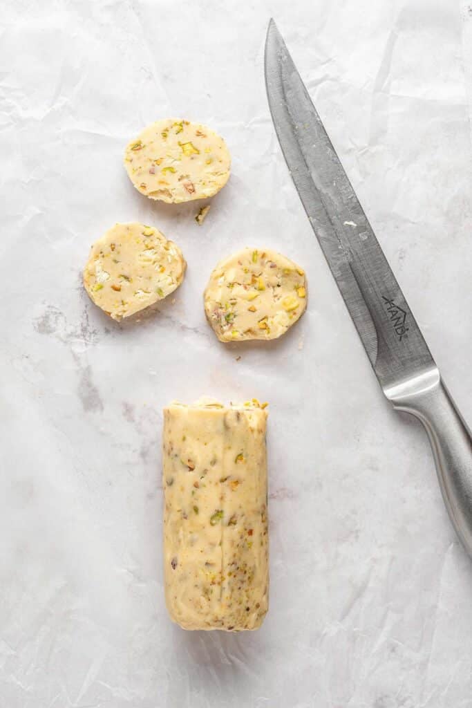 A log of pistachio shortbread dough, partially sliced, next to a large kitchen knife on a white surface.