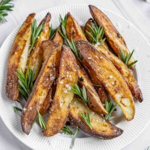 A white plate of rosemary roasted potatoes garnished with fresh rosemary sprigs on a marble surface.