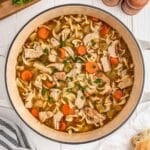 A pot of chicken noodle soup with sliced carrots, celery, chicken pieces, and pasta on a white surface.