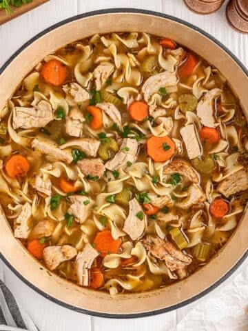 A pot of chicken noodle soup with sliced carrots, celery, chicken pieces, and pasta on a white surface.