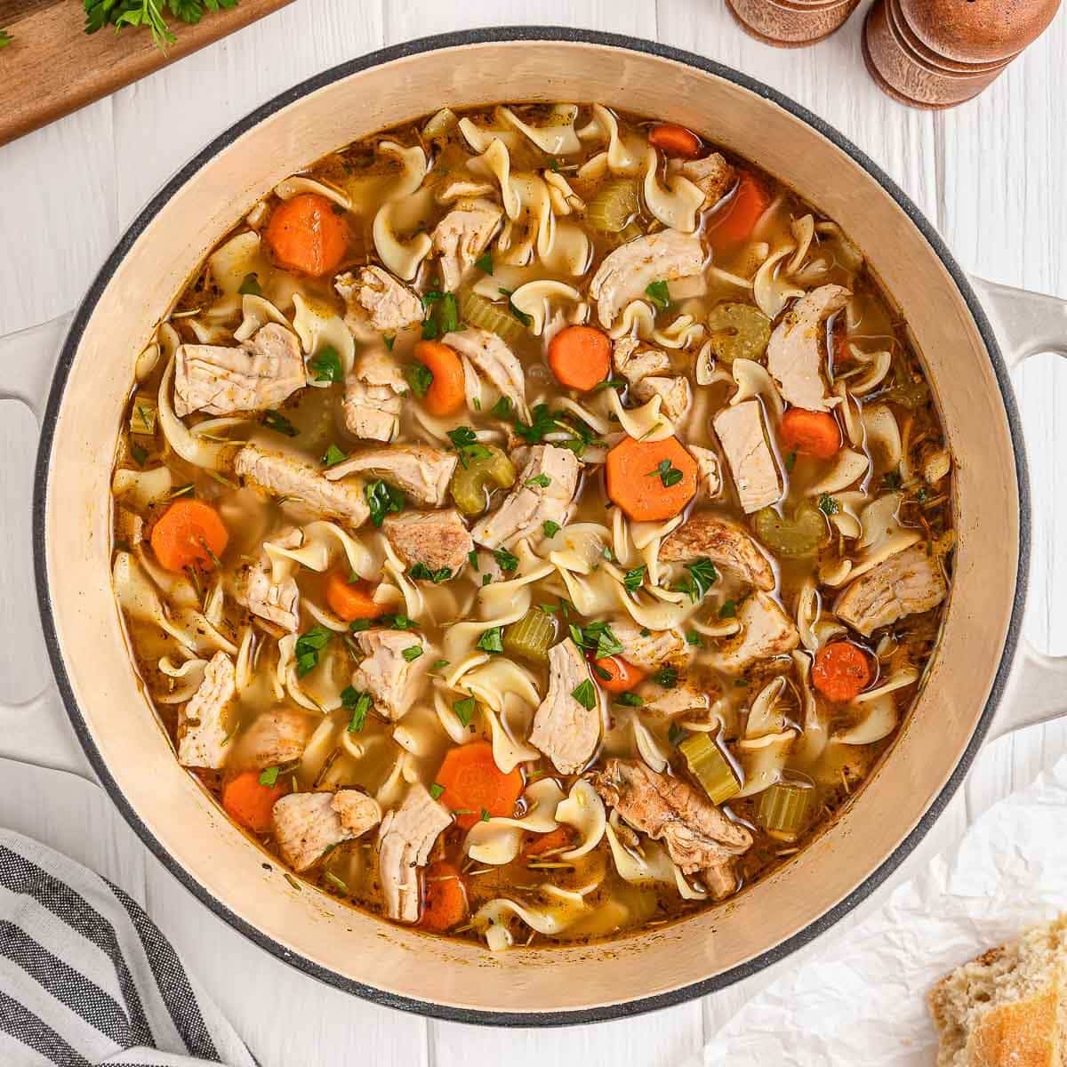 A pot of chicken noodle soup with sliced carrots, celery, chicken pieces, and pasta on a white surface.