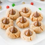 Plate of seven peanut butter blossom cookies with chocolate drops on top, surrounded by a glass of milk and festive decorations.