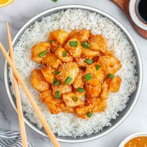 A bowl of air fryer orange chicken overtop of white rice garnished with sesame seeds and green onions.