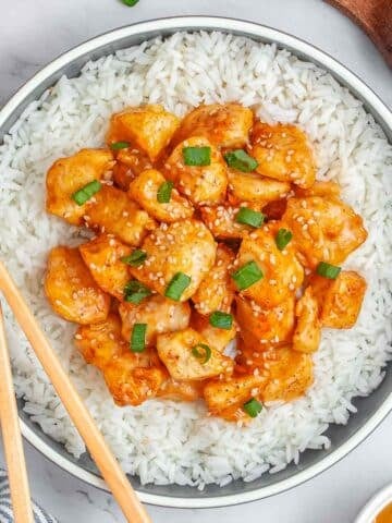 A bowl of air fryer orange chicken overtop of white rice garnished with sesame seeds and green onions.