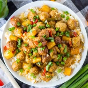A bowl of rice topped with hawaiian meatballs, diced yellow and red bell peppers, green onions, and pineapple chunks on a checkered cloth.