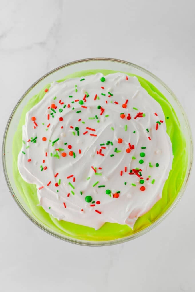 A bowl of Christmas trifle recipe with green pudding topped with whipped cream and red, green, and white sprinkles, on a white surface.