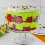A trifle dessert in a glass bowl with layers of red velvet cake, green pudding, whipped cream, and colorful sprinkles on top.