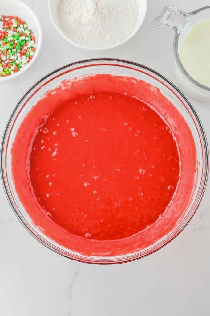 A bowl of red cake batter on a countertop surrounded by bowls of sprinkles, flour, and a measuring cup with milk.