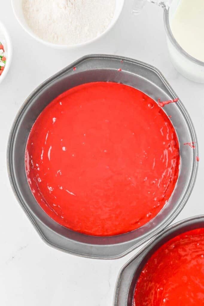 Baking pans filled with red cake batter sit on a white countertop with bowls of flour, milk, and colorful sprinkles nearby.