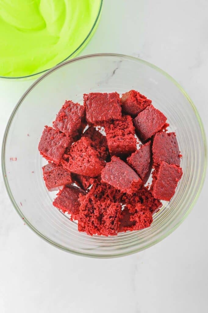A glass bowl filled with chunks of red velvet cake on a white surface. Another bowl with green frosting is partially visible in the background.