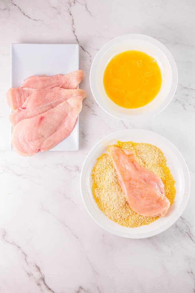 Three chicken breasts on a white plate, next to a bowl of beaten eggs and a plate of breaded chicken breasts.