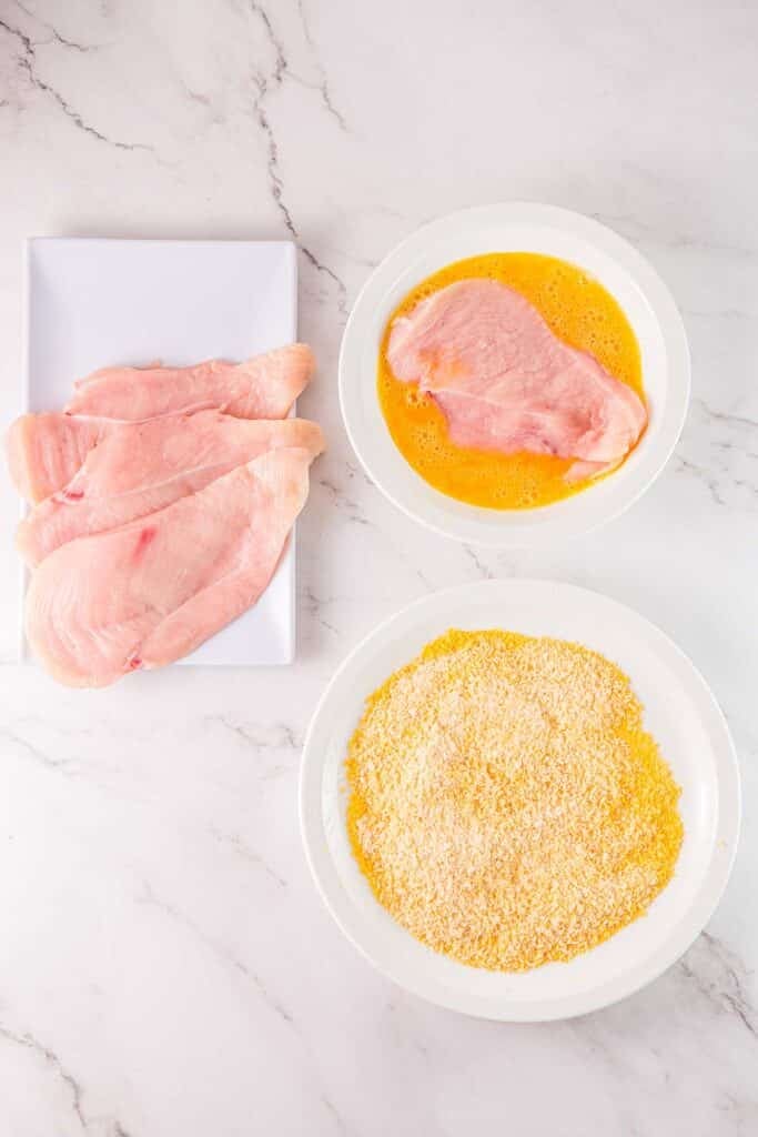 Raw chicken breasts on a plate, with one cutlet dipped in beaten eggs, and a bowl of breadcrumbs nearby.