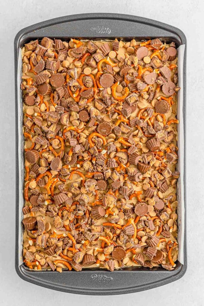 Baking tray filled peanut butter chips, mini peanut butter cups and pretzels covering the peanut butter frosting layer.