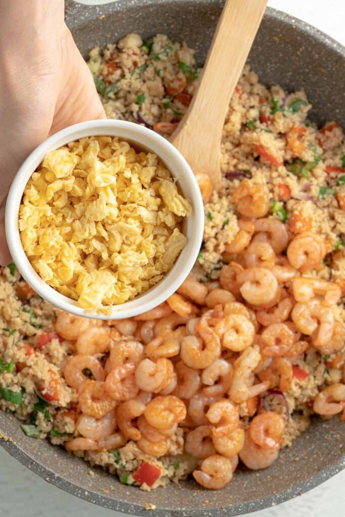 A hand holds a small bowl of scrambled eggs above a pan containing riced cauliflower, shrimp, and vegetables.