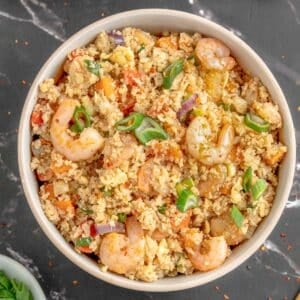 Bowl of shrimp cauliflower fried rice with diced vegetables and green onions on top, placed on a black marble surface.