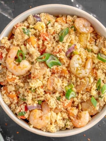 Bowl of shrimp cauliflower fried rice with diced vegetables and green onions on top, placed on a black marble surface.