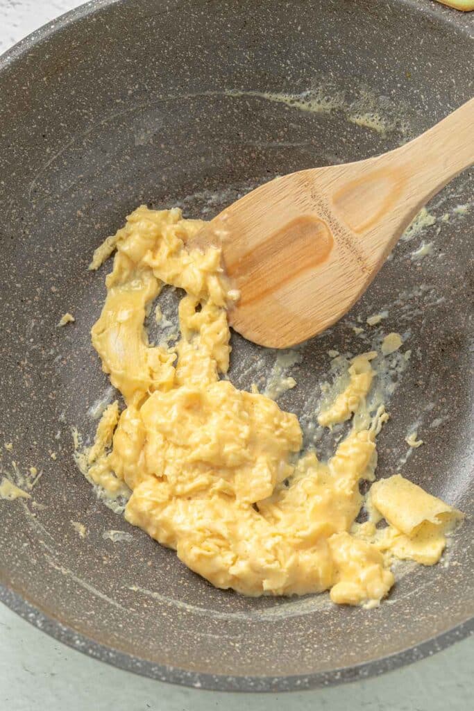 Scrambled eggs being cooked in a non-stick frying pan with a wooden spatula.
