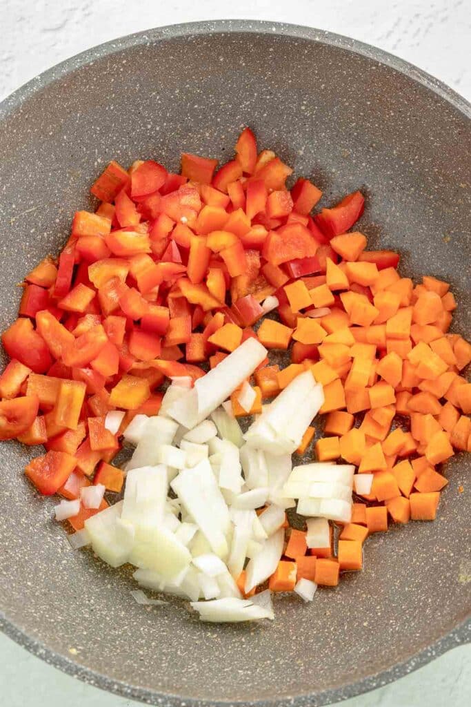 Chopped red bell pepper, carrots, and onions in a gray frying pan.