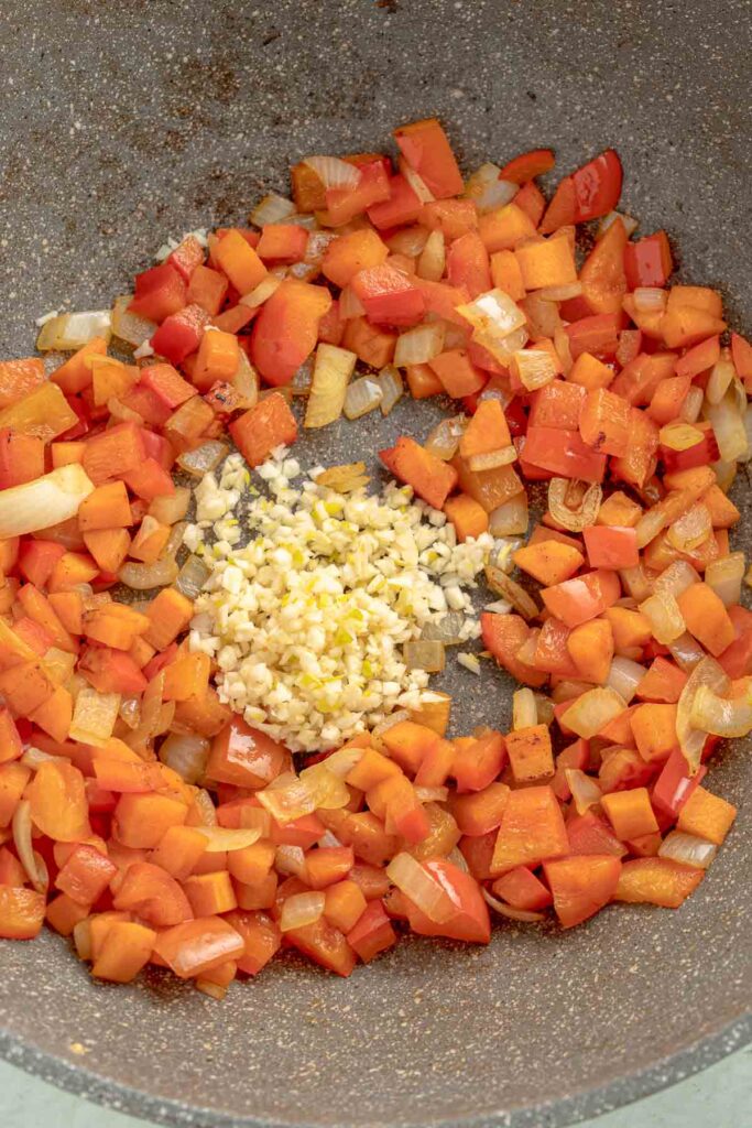 Chopped onions and red peppers sautéing in a pan with minced garlic in the center.