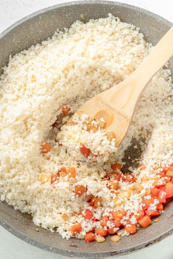 A pan containing cauliflower rice with red bell pepper pieces, stirred with a wooden spatula.