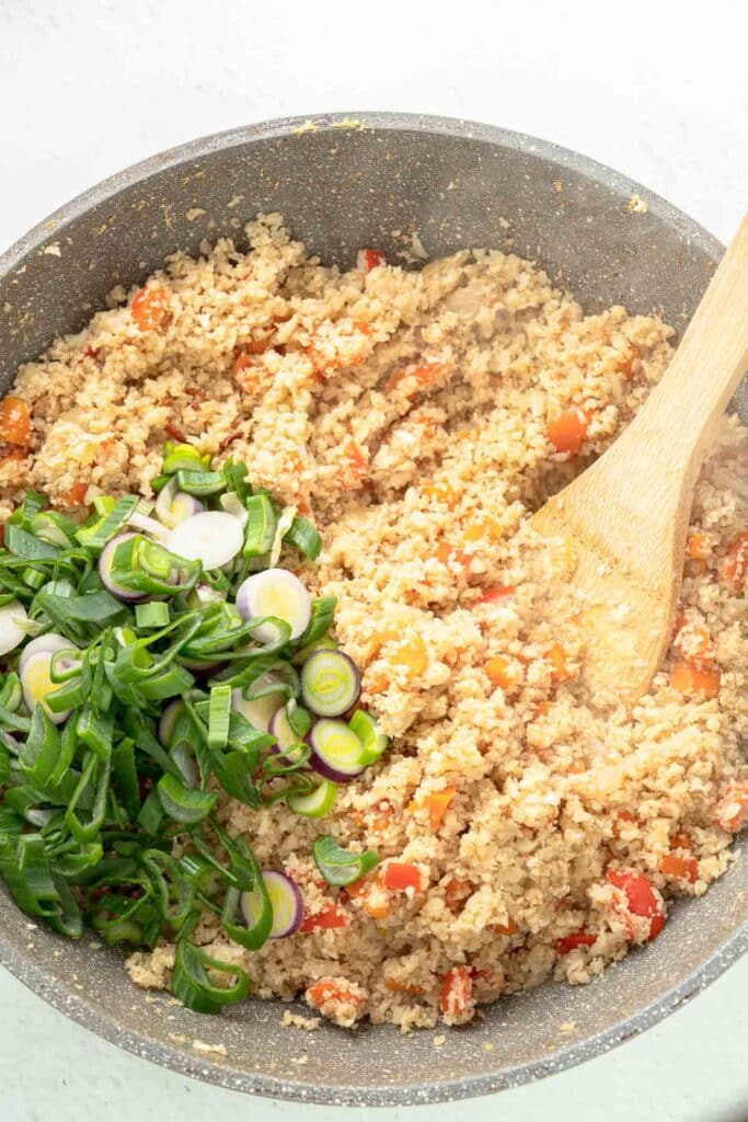 A skillet with cooked cauliflower rice mixed with diced red peppers and a wooden spoon, topped with sliced green onions.
