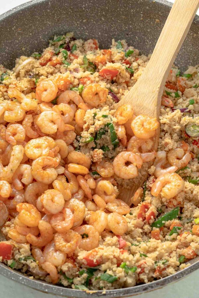 A skillet filled with shrimp, cauliflower rice, and vegetables, being stirred with a wooden spoon.