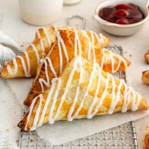 three cherry turnovers with icing drizzle, stacked on a cooling rack, with a bowl of cherry filling in the background.