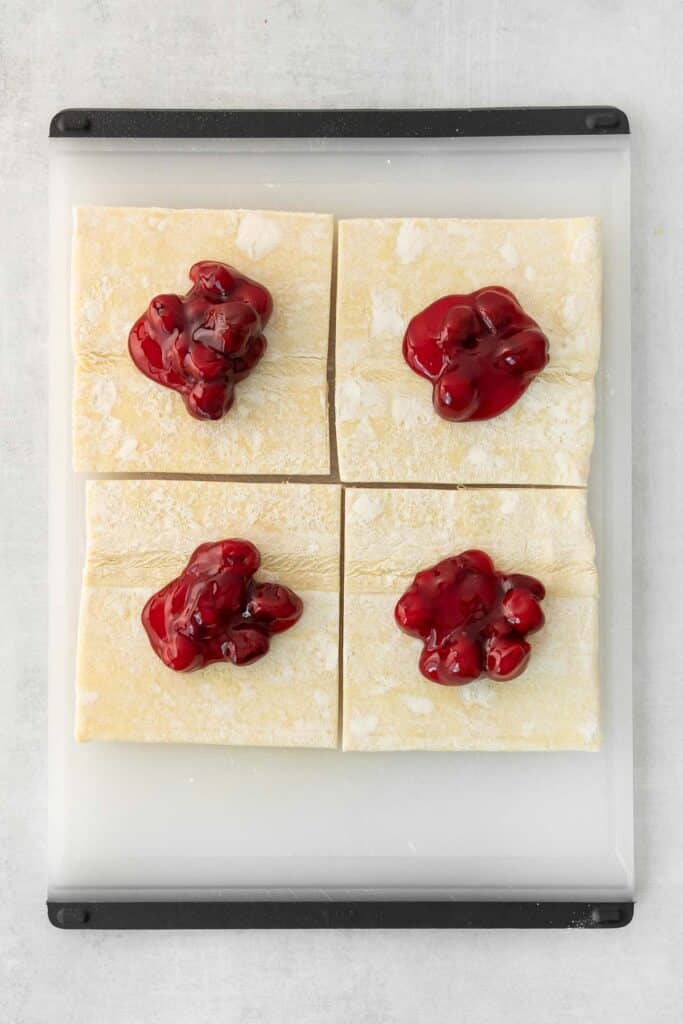 Four square pastries on a white cutting board, each topped with cherry filling.