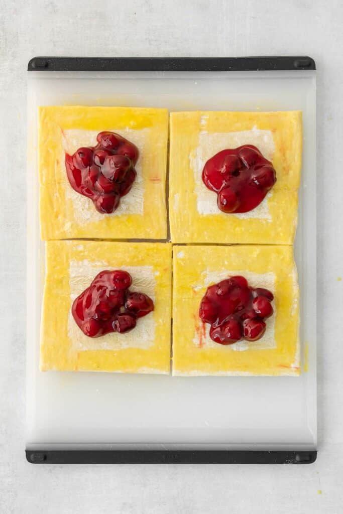 Four square pastry dough pieces on a tray, each topped with a dollop of cherry pie filling.