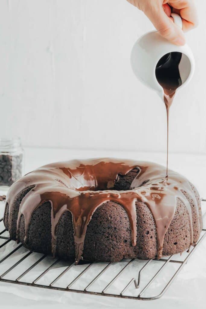 Chocolate glaze is being poured over the cake on a cooling rack.