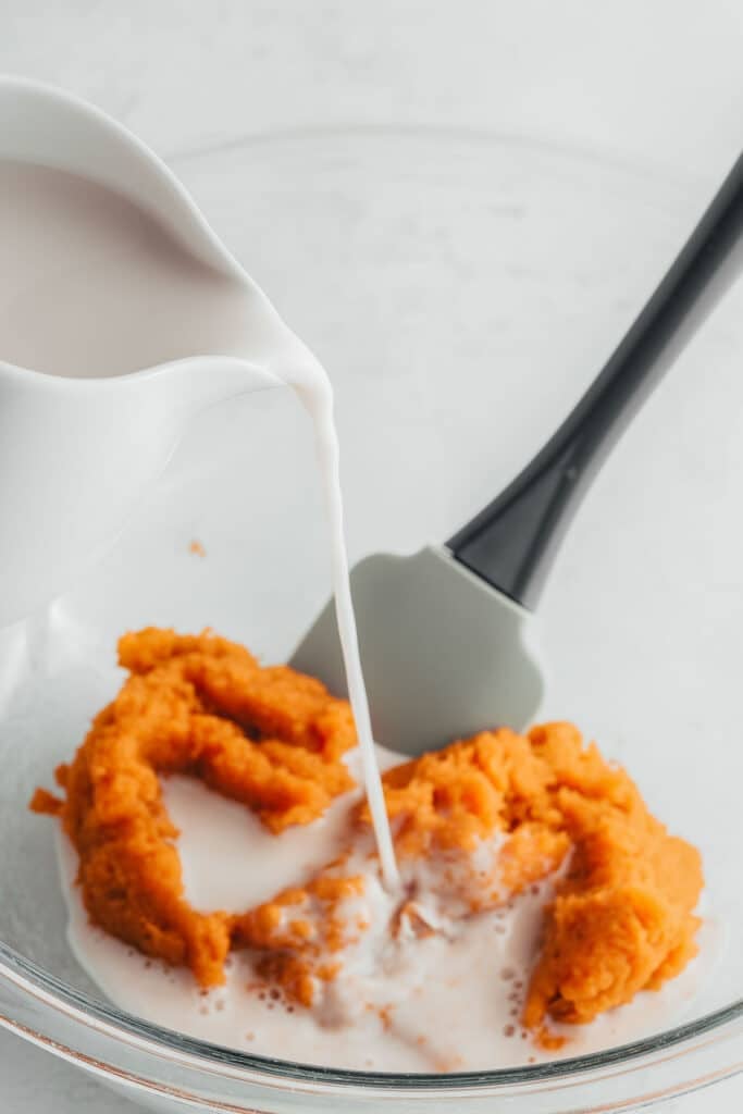 Almond milk being poured into a bowl of sweet potato puree, with a spatula resting in the mixture.