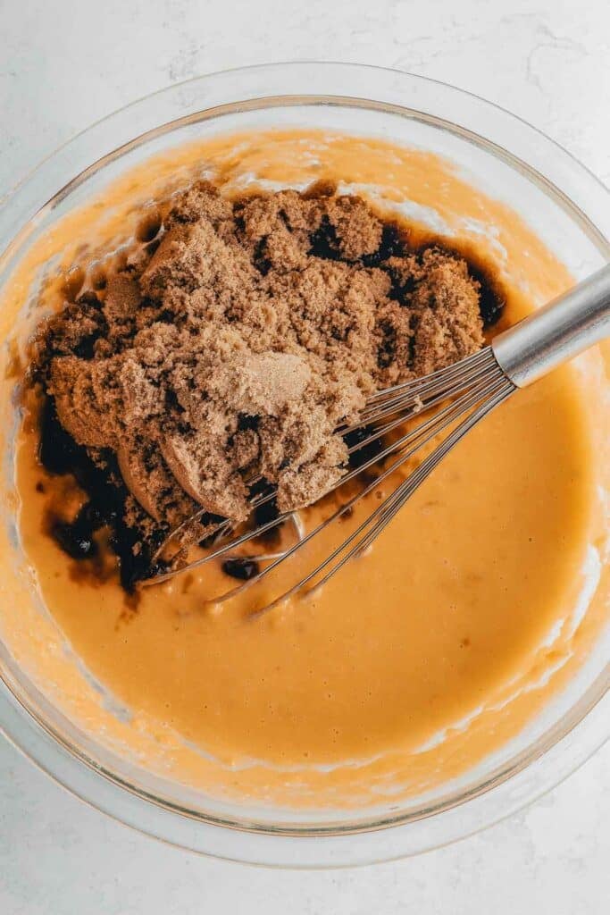 Glass bowl containing a mixture of brown sugar, vanilla, and apple cider vinegar added to sweet potato puree and almond milk with a wire whisk on a white surface.