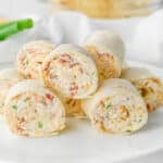 Several rolled crack chicken pinwheels made with tortillas filled with cream cheese, chicken, bacon chopped green onion, and herbs, stacked on a white plate.