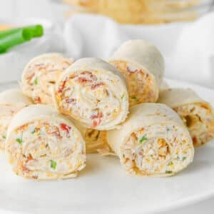 Several rolled crack chicken pinwheels made with tortillas filled with cream cheese, chicken, bacon chopped green onion, and herbs, stacked on a white plate.