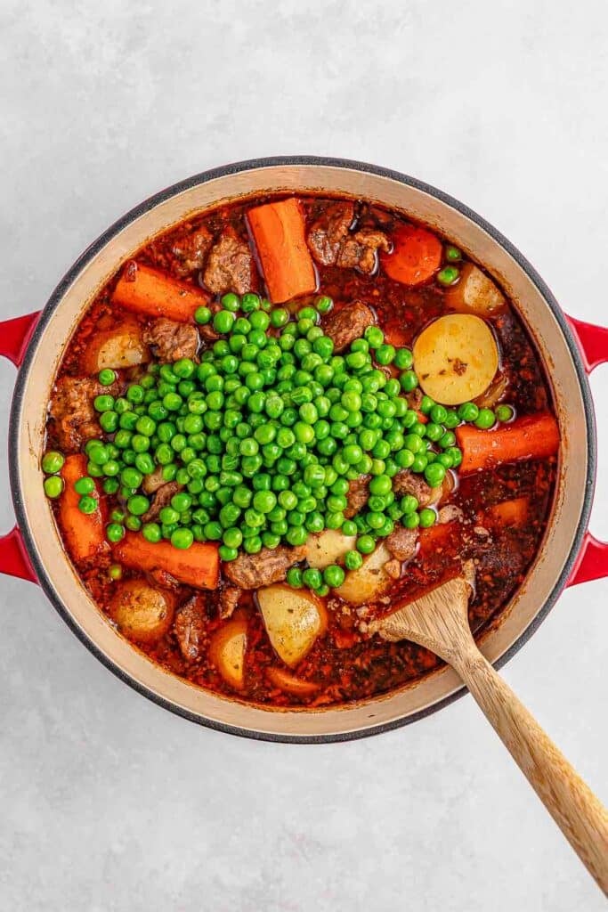 A pot of beef stew with carrots, potatoes, and peas on top ready to be mixed with a wooden spoon.
