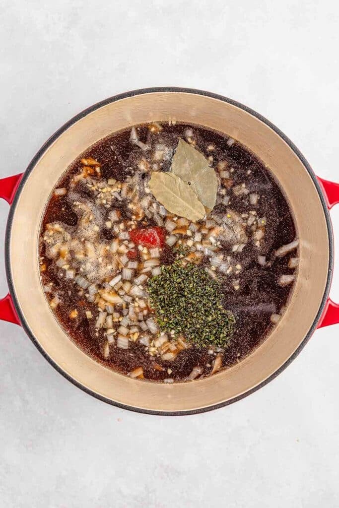 A red pot filled with broth, tomato paste, chopped onions, spices, bay leaves, and a sprig of herbs, viewed from above.