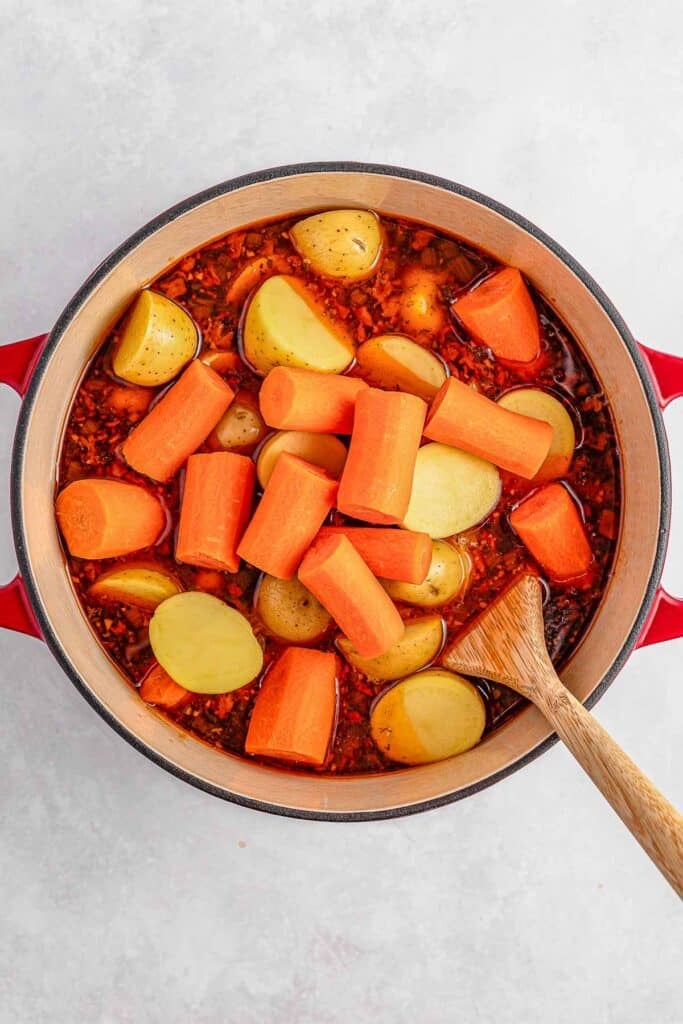 A pot of meat filled with chunks of carrots and potatoes in a beef broth, stirred with a wooden spoon.