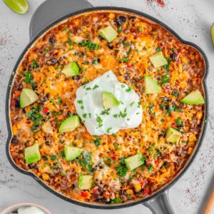 A skillet filled with mexican beef and rice topped with melted cheese, avocado, and a dollop of sour cream.