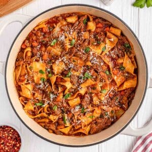 A pot of short rib ragu pappardelle pasta with shredded beef sort ribs, vegetables, and tomato sauce, garnished with grated cheese and herbs, on a white wooden surface.