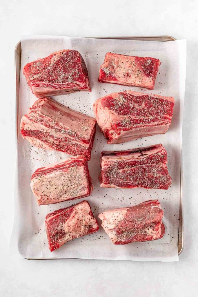 Eight raw, seasoned beef short ribs on a parchment-lined baking sheet.