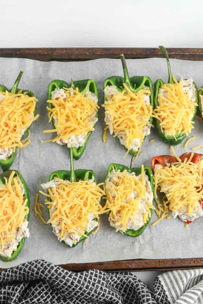 Green poblano pepper halves filled with a cream cheese mixture and topped with shredded cheese are arranged on a baking sheet with parchment paper. A black and white checkered cloth is partially visible.