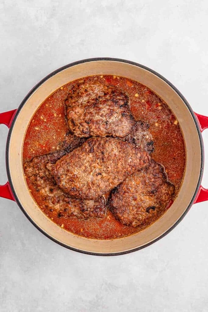 A red pot filled with cooked steaks in a red sauce.